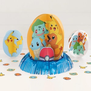 May include: A collection of colorful Pokémon-themed party decorations. The centerpiece is an orange egg-shaped decoration featuring Pikachu, Bulbasaur, Squirtle, and Charmander. Additional decorations include blue and white paper cutouts of Pokémon characters and confetti.