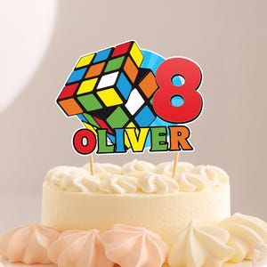 May include: A birthday cake with a topper featuring a colorful Rubik's Cube design, the number 8 in red, and the name "Oliver" in colorful letters. The cake is decorated with white frosting and meringue cookies.