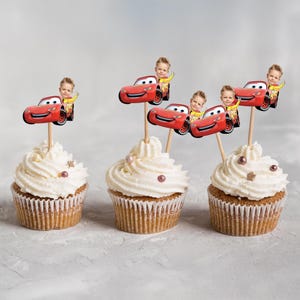 May include: Three cupcakes with white frosting, each topped with a red race car and a child's image. The cupcakes are in brown paper liners and decorated with small pink and gold sprinkles. The background is a light gray.