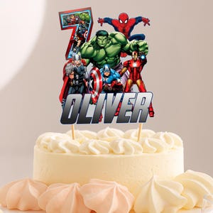 May include: A cake topper featuring the Avengers characters, including Hulk, Iron Man, and Spider-Man, with the name "OLIVER" and the number "7". The topper is placed on a white frosted cake with meringue cookies.
