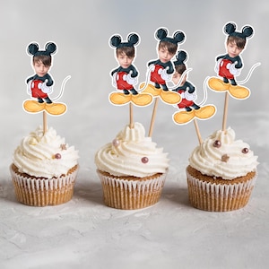 May include: Three cupcakes with white frosting and decorative sprinkles. Each cupcake is topped with a Mickey Mouse-themed cake topper featuring a child's face. The toppers are on wooden sticks.