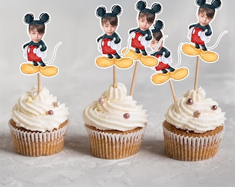 May include: Three cupcakes with white frosting and decorative sprinkles. Each cupcake is topped with a Mickey Mouse-themed cake topper featuring a child's face. The toppers are on wooden sticks.
