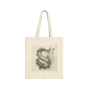 Può includere: Borsa tote color crema con una lettera "S" decorativa con un design a farfalla. La lettera è turchese con accenti dorati e ornata di fiori blu. La farfalla ha ali bianche e giallo pallido.