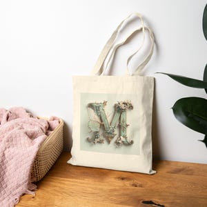 Könnte beinhalten: Eine cremefarbene Tragetasche mit einer detaillierten Illustration des Buchstabens "M". Der Buchstabe ist mit floralen und Schmetterlingsakzenten in Grün-, Blau- und Cremetönen verziert. Die Tasche hat lange Henkel und ist aus robustem Stoff gefertigt.