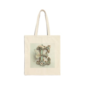 Può includere: Borsa tote color crema con un design dettagliato della lettera "E". La lettera è decorata con una farfalla e accenti floreali nei toni del blu, verde e crema. La borsa ha un design rettangolare con due manici.