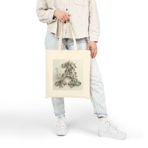 Può includere: Borsa tote color crema con la lettera "A" decorativa, una farfalla e dettagli floreali. La lettera è in stile vintage, con una palette di colori azzurro chiaro e oro. La borsa è realizzata in tessuto resistente e ha manici lunghi.