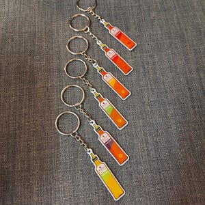 May include: Keyrings with bottle-shaped charms in a variety of colours, including orange, yellow, and green. Each charm has a silver chain and ring. The bottle designs have a white border and are arranged on a grey surface.