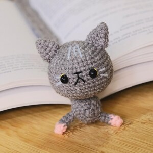Puede incluir: Marcapáginas de gato gris tejido a ganchillo con patas rosas y ojos negros. La cabeza del gato es redonda con orejas puntiagudas y detalles blancos. El marcapáginas está colocado sobre un libro abierto, sobre una superficie de madera.