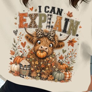 Könnte beinhalten: Ein cremefarbenes Sweatshirt mit Rundhalsausschnitt und einer Grafik eines Hochlandrindes mit einer Schleife im Leopardenmuster. Das Rind hält einen kleinen Kürbis im Maul, umgeben von Herbstblättern, Kürbissen und einem Kaffeebecher. Der Text lautet I CAN EXPLAIN.