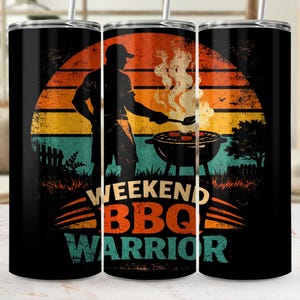 May include: A black tumbler with a graphic of a person grilling at a barbecue. The design features a sunset in orange, yellow, and teal, with the words "WEEKEND BBQ WARRIOR" in white, orange, and teal.
