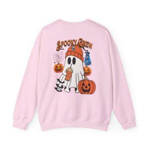 Peut inclure: Un sweat-shirt rose clair avec un motif sur le thème d'Halloween. Le dessin représente un fantôme portant un bonnet orange, tenant une boisson et entouré de citrouilles et de chauves-souris. Les mots "Spooky Crew" sont imprimés au-dessus du fantôme.
