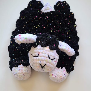 May include: A black and white crocheted lamb with colourful flecks throughout the yarn. The lamb is lying on its side with its eyes closed. The text "@madebykarla" is visible in the bottom left corner of the image.