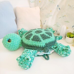 Op de afbeelding: Een gehaakt groen knuffeldier in de vorm van een schildpad met een grote schil en &eacute;&eacute;n oog. De schildpad ligt op een wit oppervlak.