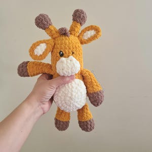 Puede incluir: Un juguete de jirafa de ganchillo hecho a mano en tonos amarillos, marrones y blancos. La jirafa tiene un ojo guiñado y está siendo sostenida. El texto "@madebykarla" es visible en el lateral.