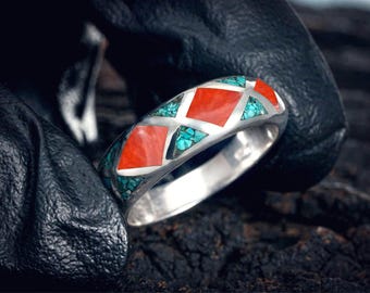 May include: A silver ring with a geometric design featuring red and turquoise inlays. The ring's band is polished silver, and the inlays are arranged in a repeating pattern. The ring is shown against a dark background.