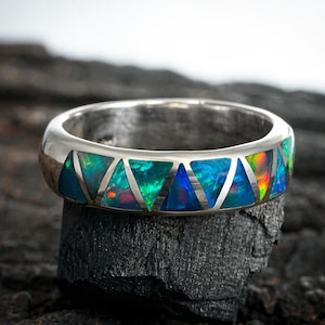 May include: A silver ring featuring a band of triangular opal stones. The opals exhibit a spectrum of colours, including blue, green, and red. The ring is set against a dark, textured backdrop.