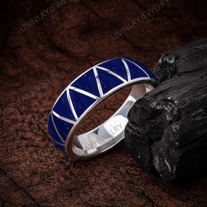 May include: A silver ring with a geometric design. The ring has a band of triangular blue inlays set within a silver frame. The ring is displayed on a dark brown surface next to a piece of black charcoal.