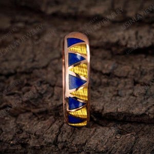 May include: A rose gold ring with a geometric design. The ring features alternating blue triangular inlays and yellow, wood-grain-textured inlays. The ring is set against a dark, textured background.