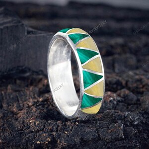 May include: A silver ring featuring a geometric pattern of alternating green and yellow triangular inlays. The ring's design is set against a dark, textured backdrop, emphasizing the vibrant colours and intricate pattern of the jewellery.