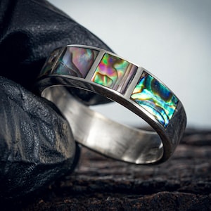 Könnte beinhalten: Ein silberner Ring mit drei rechteckigen Einlagen aus schillerndem Abalone-Muschelmaterial. Das Abalone zeigt eine Farbpalette von Grün, Rosa und Lila. Der Ring ist vor einem dunklen Hintergrund platziert.