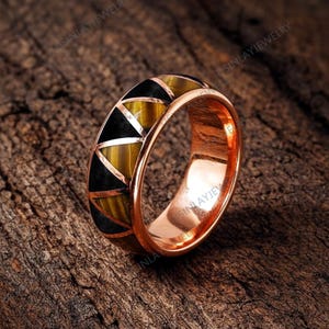 May include: A rose gold ring with a geometric design. The ring features alternating black and tiger's eye-colored triangular inlays around the band. The ring is set against a dark wood grain background.