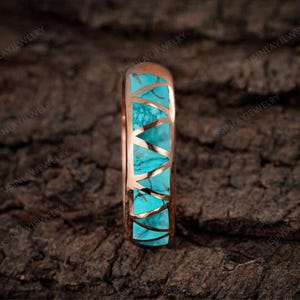 May include: A rose gold ring with a turquoise inlay. The turquoise is cut into geometric shapes and set within the ring's band. The ring is displayed on a dark, textured surface.