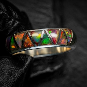 May include: Silver ring with a band featuring triangular inlays of vibrant, multicoloured opal. The opal displays a spectrum of colours, including orange, green, and red, set against a dark background. The ring's design is modern and eye-catching.