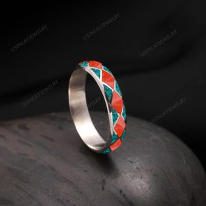 May include: A silver ring with a geometric pattern of red and turquoise inlays. The ring is set against a dark background, highlighting the bright colors of the inlaid design. The ring is a band style, with the text "USINLAYJEWELRY" visible.