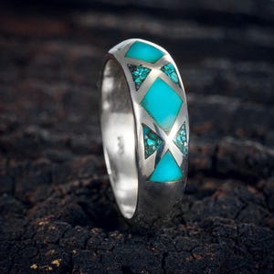 Puede incluir: Un anillo de plata con incrustaciones de turquesa. El anillo presenta un patrón repetitivo de diamantes y triángulos turquesas. El anillo está sobre un fondo oscuro y texturizado.