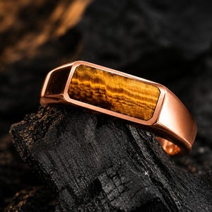 Puede incluir: Un anillo en tono oro rosa con una incrustación rectangular de piedra ojo de tigre en tonos marrones y dorados. La banda tiene un acabado metálico pulido y una superficie superior plana que enmarca el patrón rayado natural de la piedra.