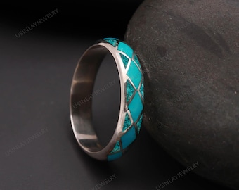 Könnte beinhalten: Ein silberner Ring mit türkisfarbener Einlage in einem Rautenmuster. Der Ring ist vor einem dunklen Hintergrund mit einem großen, dunkelgrauen Stein platziert. Das geometrische Design kombiniert Silber und leuchtendes Türkis.