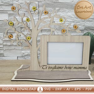 Può includere: Cornice in legno a forma di albero, decorata con fiori gialli e arancioni. La cornice ha un'apertura rettangolare per una foto e il testo italiano "Ti vogliamo bene mamma". Realizzata in legno marrone chiaro.