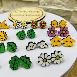 May include: A collection of wooden stud earrings in various shapes, including flowers, leaves, clouds, stars, and suns. The earrings are painted in bright colors like yellow, green, pink, and white. The earrings are displayed on a white surface.