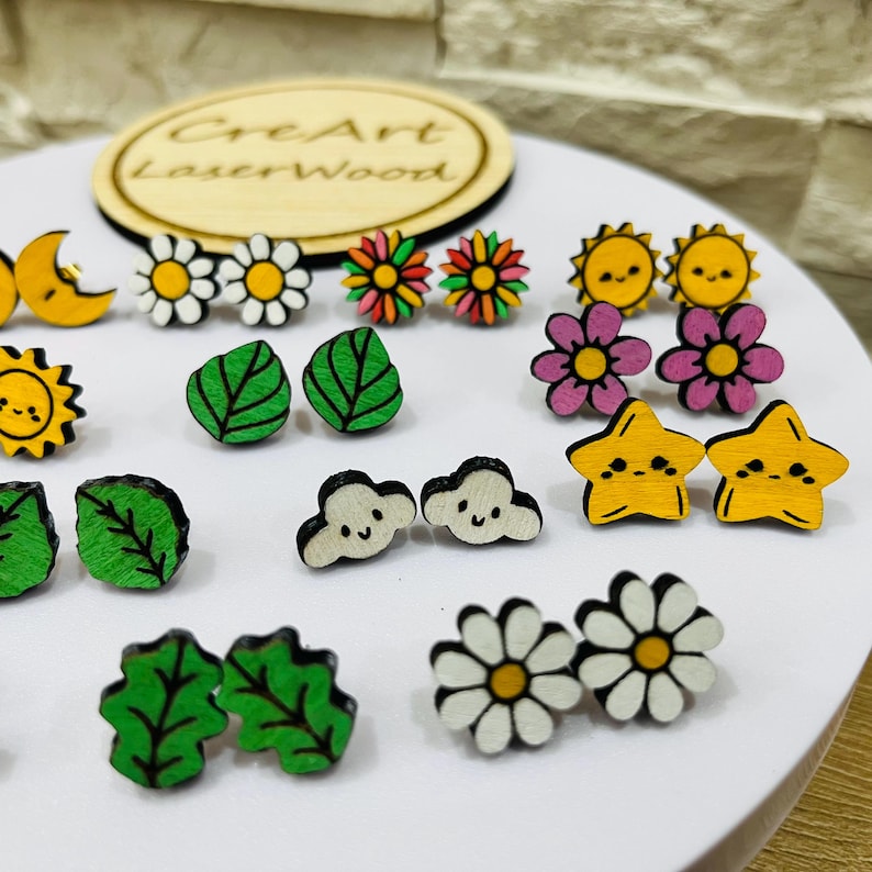 May include: A collection of wooden stud earrings in various shapes, including flowers, leaves, clouds, stars, and suns. The earrings are painted in bright colors like yellow, green, pink, and white. The earrings are displayed on a white surface.