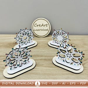 Può includere: Quattro ornamenti a forma di ruota panoramica in legno tagliato al laser con luci colorate. Ogni ornamento è bianco con una base nera e poggia su una base ovale in legno. Sullo sfondo un cerchio di legno con la scritta "CreArt LaserWood".