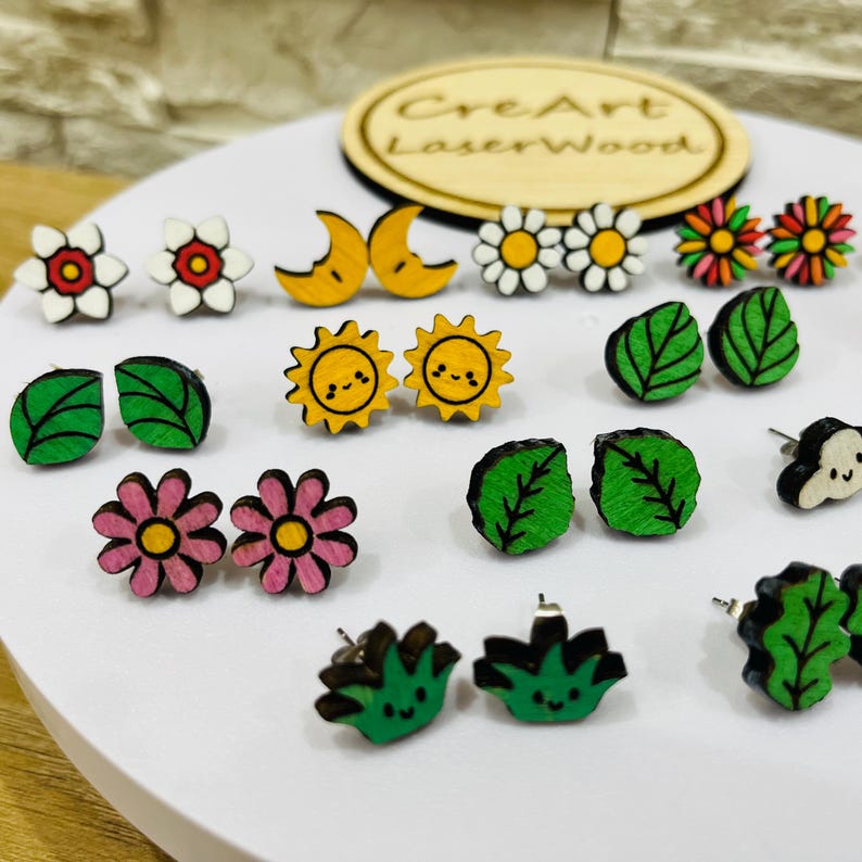 May include: A collection of wooden stud earrings in various designs, including flowers, leaves, suns, moons, and daisies. The earrings are painted in bright colors like pink, yellow, green, and white. The earrings are displayed on a white surface.