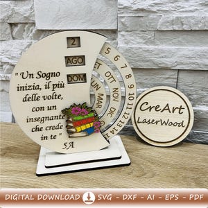 Può includere: Un calendario da scrivania in legno con un design circolare, con la data '2' e il mese 'AGO'. Il calendario include una citazione in italiano e una pila di libri con fiori. È presente anche un cerchio di legno separato con il testo 'CreArt LaserWood'.