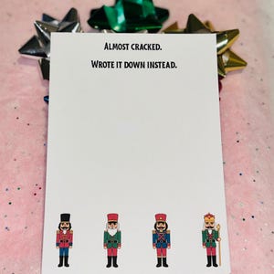 May include: A white notepad with the text "Almost cracked. Wrote it down instead." and four cartoon nutcracker soldiers. The notepad is decorated with a green, silver, and gold bow.