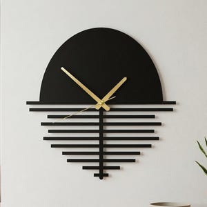 Pode incluir: Um relógio de parede preto moderno com ponteiros dourados. O relógio apresenta um topo semicircular e um design geométrico de linhas horizontais abaixo. Está montado numa parede branca, com uma prateleira de madeira e decoração em primeiro plano.