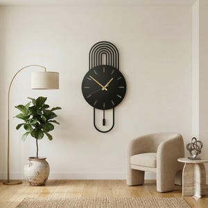 Könnte beinhalten: Eine moderne schwarze Wanduhr mit einzigartigem Design. Die Uhr hat ein rundes Zifferblatt mit goldfarbenen Zeigern und einem dekorativen, bogenförmigen oberen Teil. Sie ist an einer weißen Wand befestigt und schafft eine minimalistische Ästhetik. Ca. 60 cm hoch.