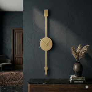 May include: A gold-colored, modern wall clock with a minimalist design. The clock features a circular face with hands, a long vertical bar extending above and below the face, and a rectangular top and arrow-shaped bottom. The clock is mounted on a dark blue wall.