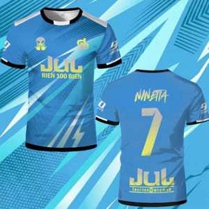 May include: A short-sleeved blue t-shirt featuring a black crew neck and sleeve trim. The front displays an alien graphic, the text JUL RIEN 100 RIEN, and a logo. The back features the name NINETTA, the number 7, and the text JUL lacrim micmak.