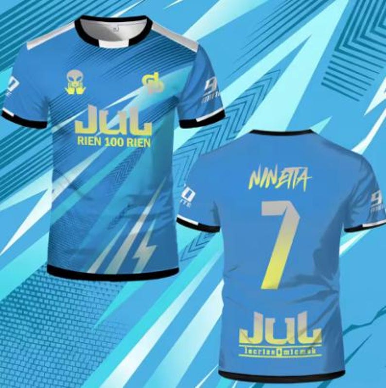 May include: A short-sleeved blue t-shirt featuring a black crew neck and sleeve trim. The front displays an alien graphic, the text JUL RIEN 100 RIEN, and a logo. The back features the name NINETTA, the number 7, and the text JUL lacrim micmak.