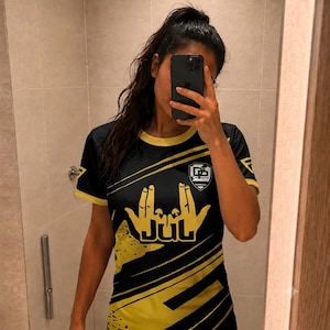May include: A black and yellow short-sleeve t-shirt featuring a graphic of two hands forming a sign above the text JUL in a bold font. The shirt includes a small shield-shaped logo on the upper chest and diagonal yellow stripes across the torso.