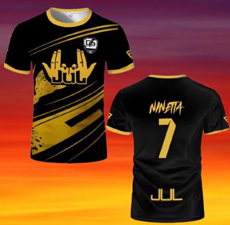 May include: A black and gold short-sleeve t-shirt featuring a graphic of hand gestures and the text JUL on the front. The back displays the name NINETTA above the number 7 and the text JUL in gold lettering, with gold trim on the collar and sleeves.