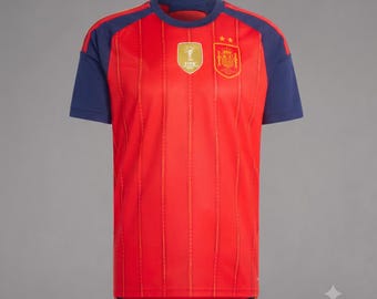 Puede incluir: Una camiseta roja de manga corta con mangas azul oscuro y cuello redondo. El frente presenta rayas verticales de color naranja, una insignia dorada de la Copa Mundial de la FIFA y el escudo de la selección nacional de fútbol de España con dos estrellas.