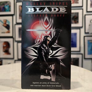 Puede incluir: Una cinta VHS negra de la película "Blade" con Wesley Snipes y Stephen Dorff. La portada presenta a un personaje sosteniendo una espada sobre un fondo rojo y blanco. El texto dice: "Contra un ejército de Inmortales, un guerrero debe derramar la primera sangre."