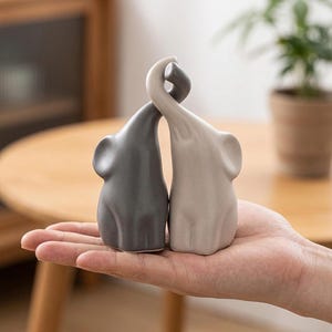 May include: Two ceramic elephant figurines, one grey and one off-white, with intertwined trunks. The elephants are displayed on a hand, with a blurred wooden table and a potted plant in the background. The figurines are approximately 7.6 cm tall.