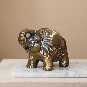May include: A bronze-colored elephant figurine with intricate detailing. The elephant has its trunk raised and is adorned with decorative patterns on its ears and body. It is placed on a white marble surface.