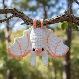 Pode incluir: Um morcego de crochê feito à mão com corpo branco e detalhes em pêssego, pendurado em um galho de árvore. O morcego tem asas grandes, orelhas pequenas e um nariz cor de pêssego. Os olhos são pretos.